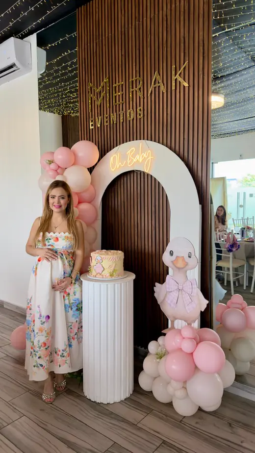 Mamá junto al pastel y a un arco de globos en tonos rosas durante un baby shower en Merak Eventos