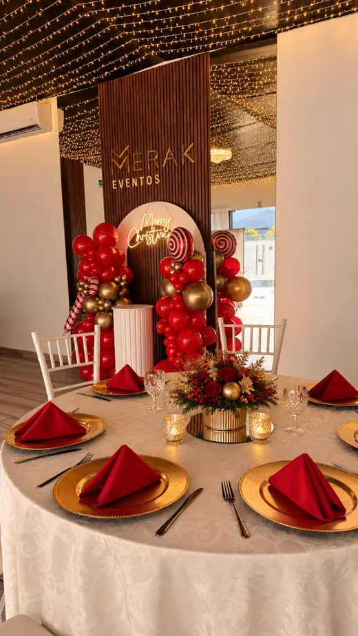 Mesa redonda montada con mantelería clara, vajilla dorada y servilletas rojas, con arco de globos al fondo, en una posada en Merak Eventos